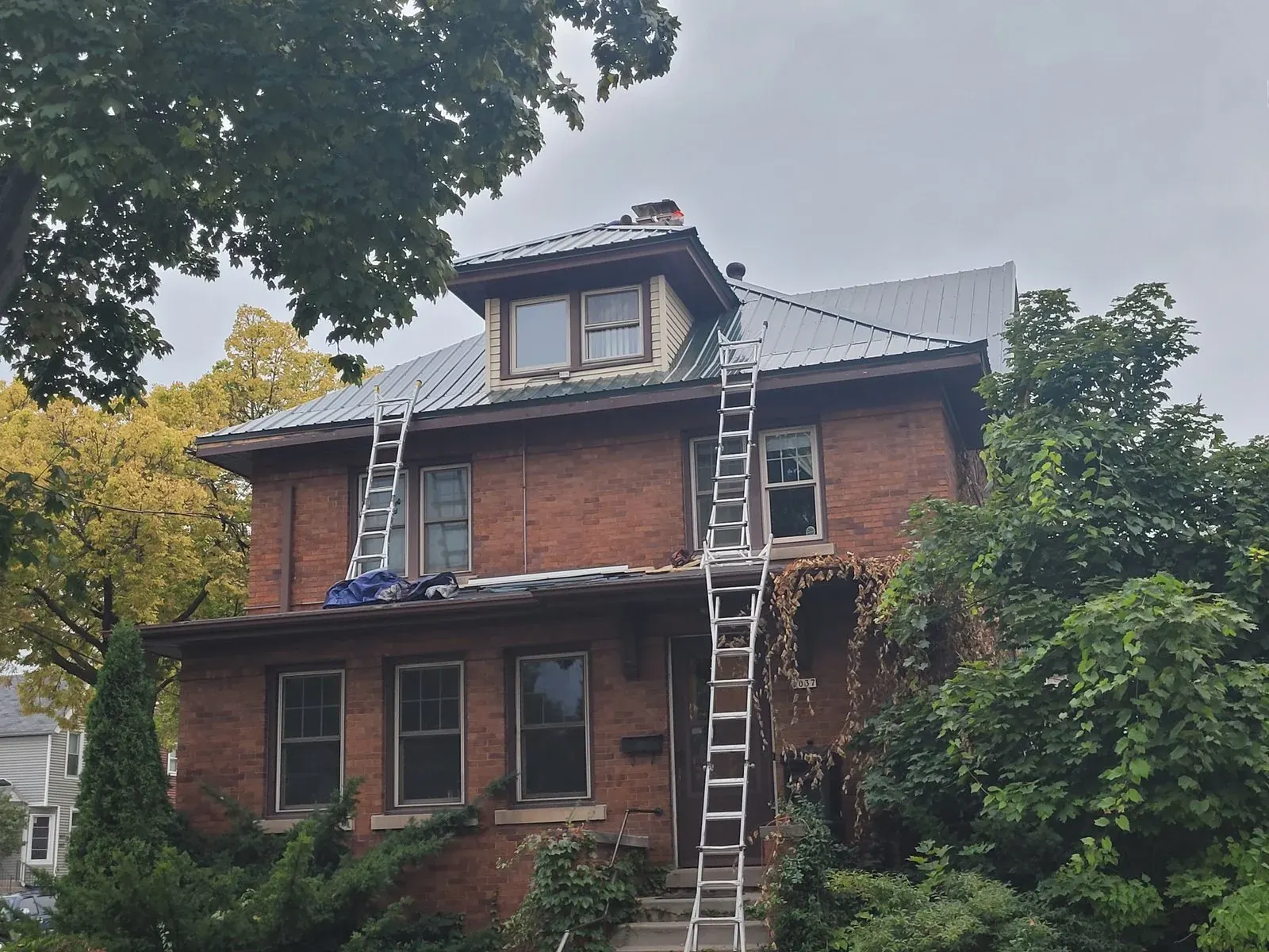Red brick house with metal roof and two ladders leaning against upper levels during renovation