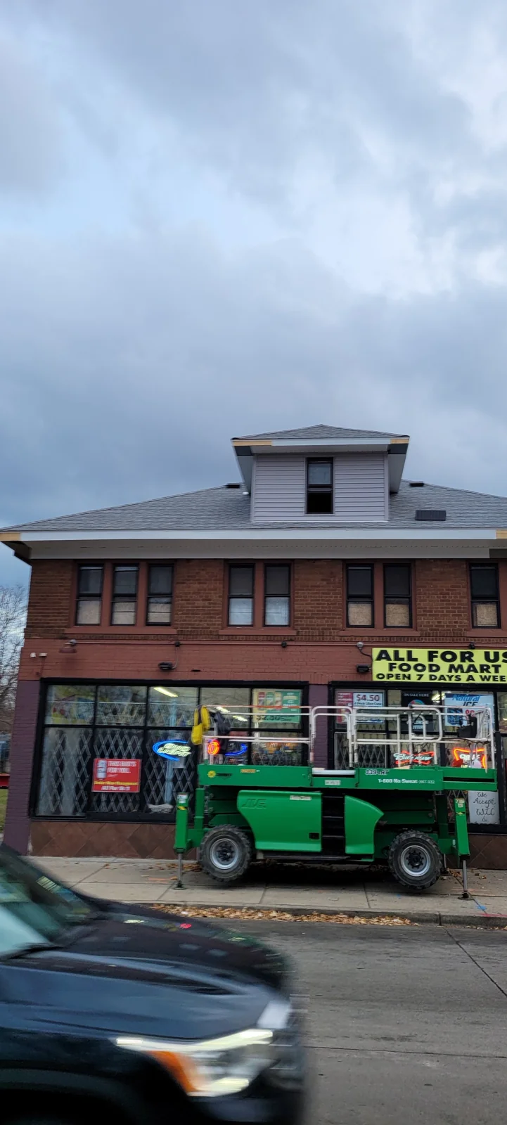 Brick building with "All for Us Food Mart" sign and green aerial lift platform parked in front