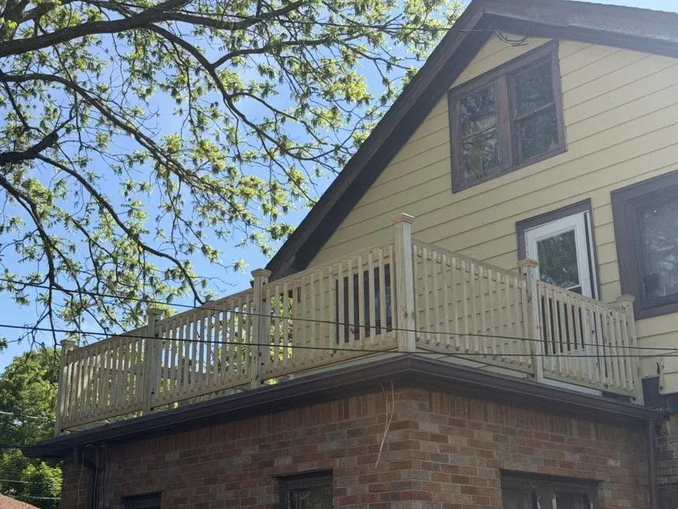 Two-story house with tan siding and wooden deck railing overlooking brick foundation