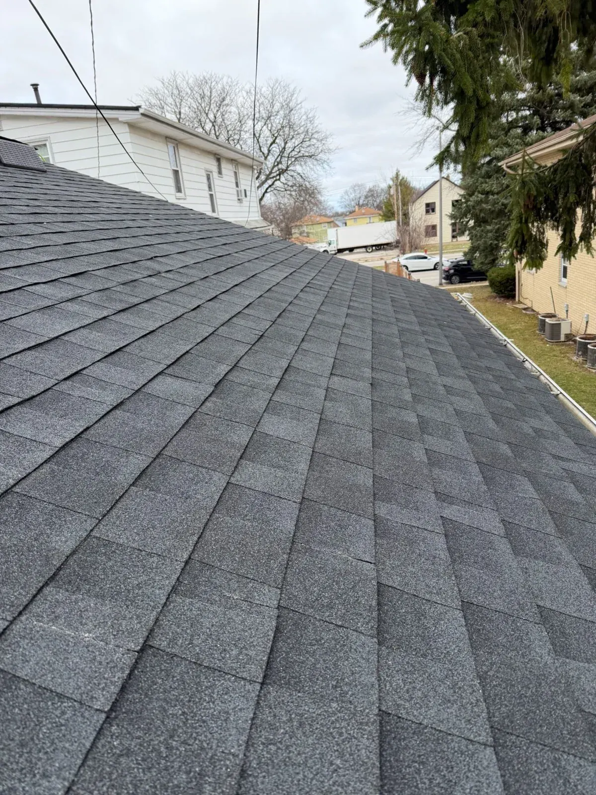 Newly installed dark gray asphalt shingles on residential roof with neighborhood houses visible in background