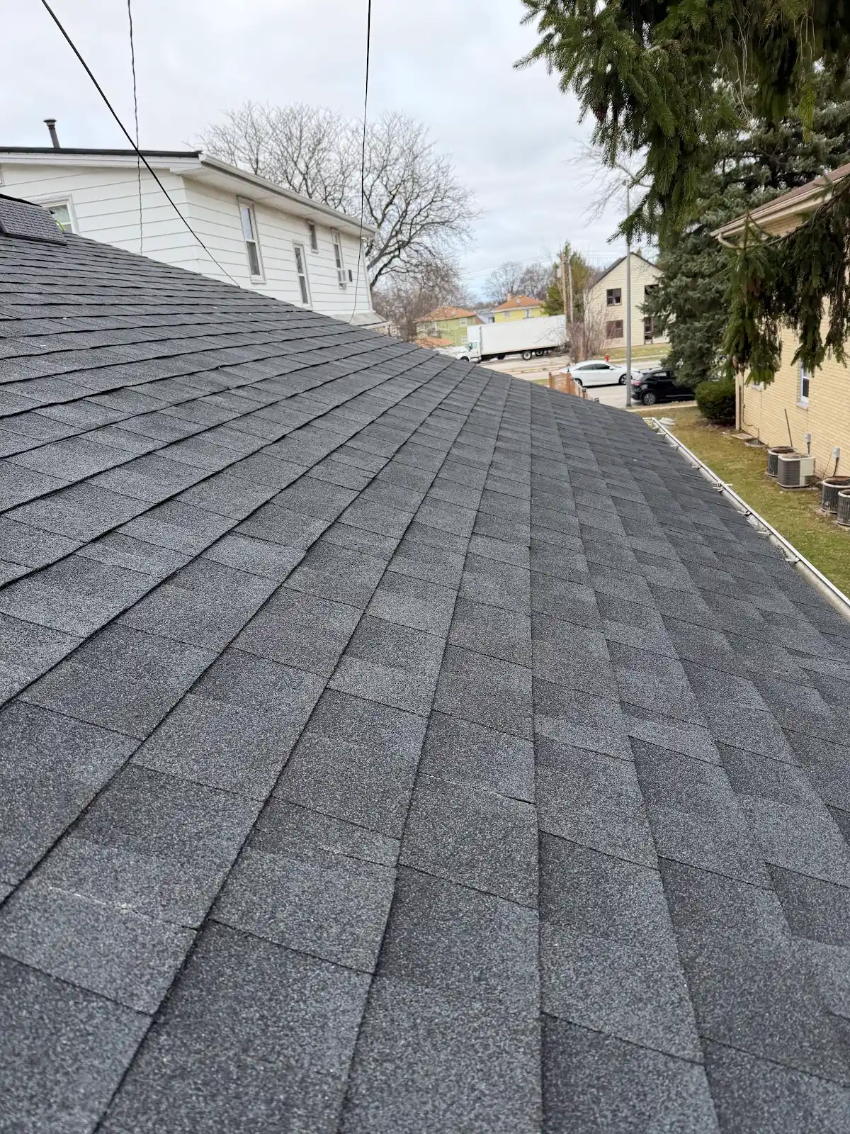 Newly installed dark gray asphalt shingles on residential roof with neighborhood houses visible in background