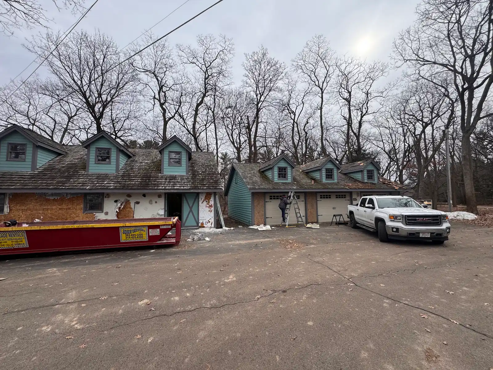 Two residential houses under renovation with boarded windows, bare trees, and a white pickup truck parked nearby