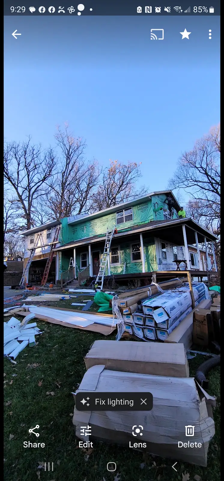 House undergoing renovation with green siding, ladders, and building materials stacked in front yard