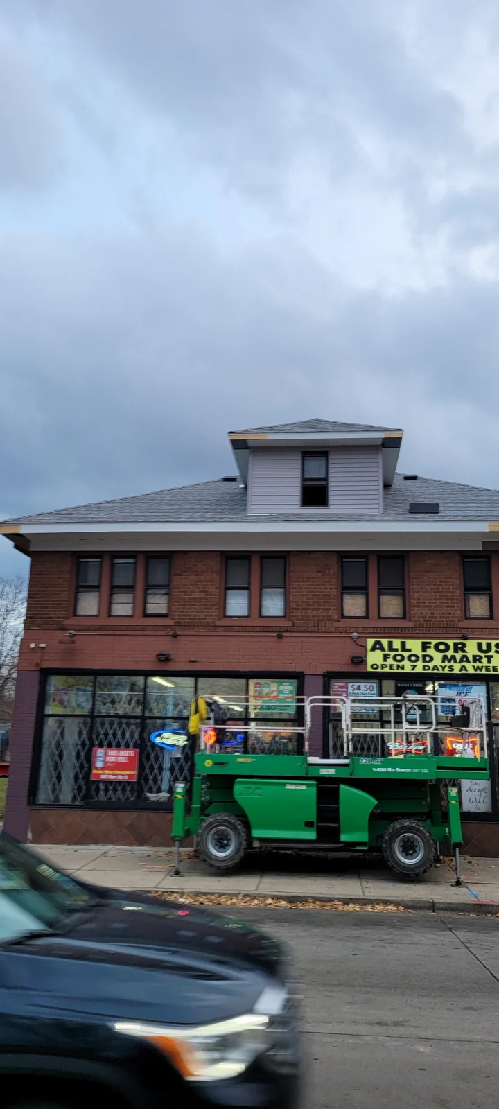Brick building with "All For Us Food Mart" storefront and green bucket lift truck parked outside